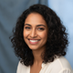 Portrait of a smiling South Asian woman, late 20s to early 30s, against a soft slate blue and white background.