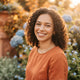 A friendly, diverse woman smiling outdoors in a sunlit setting with a soft, natural background.
